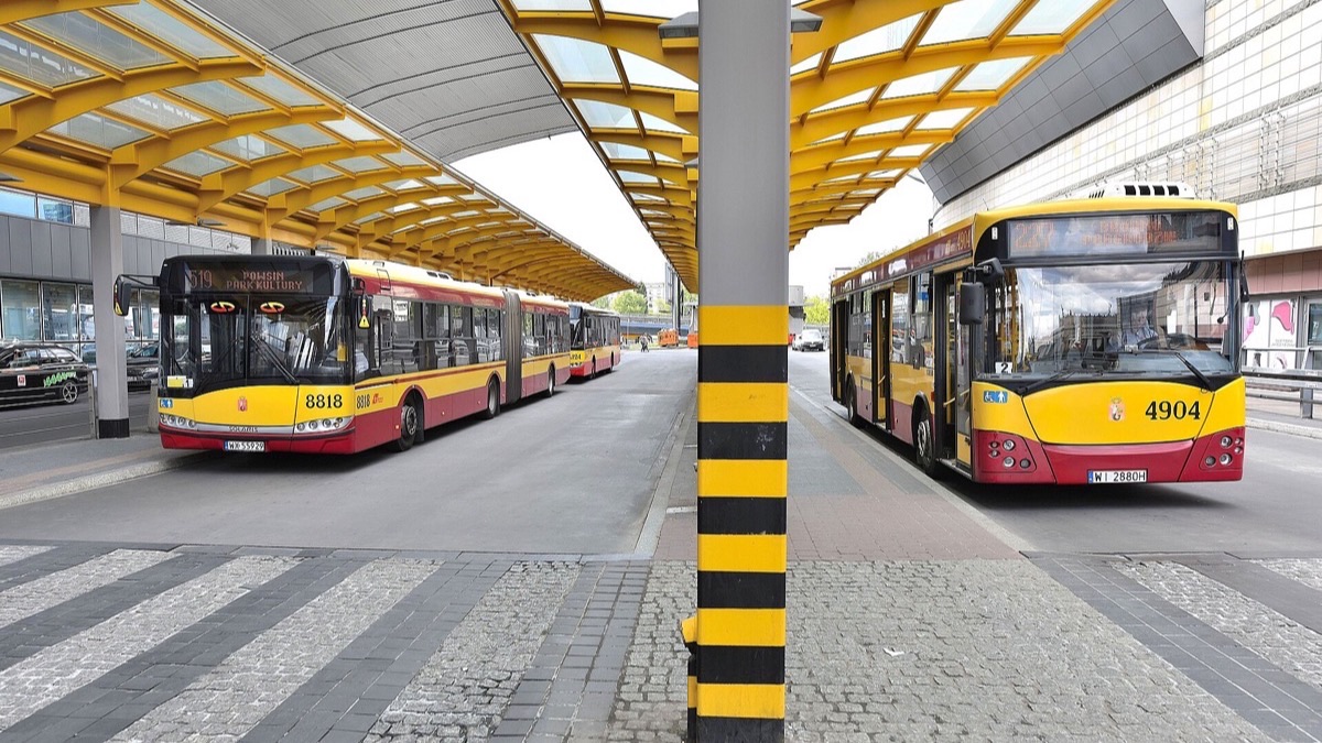Two red and yellow Warsaw city buses waiting under a modern glass canopy at a bus terminal.